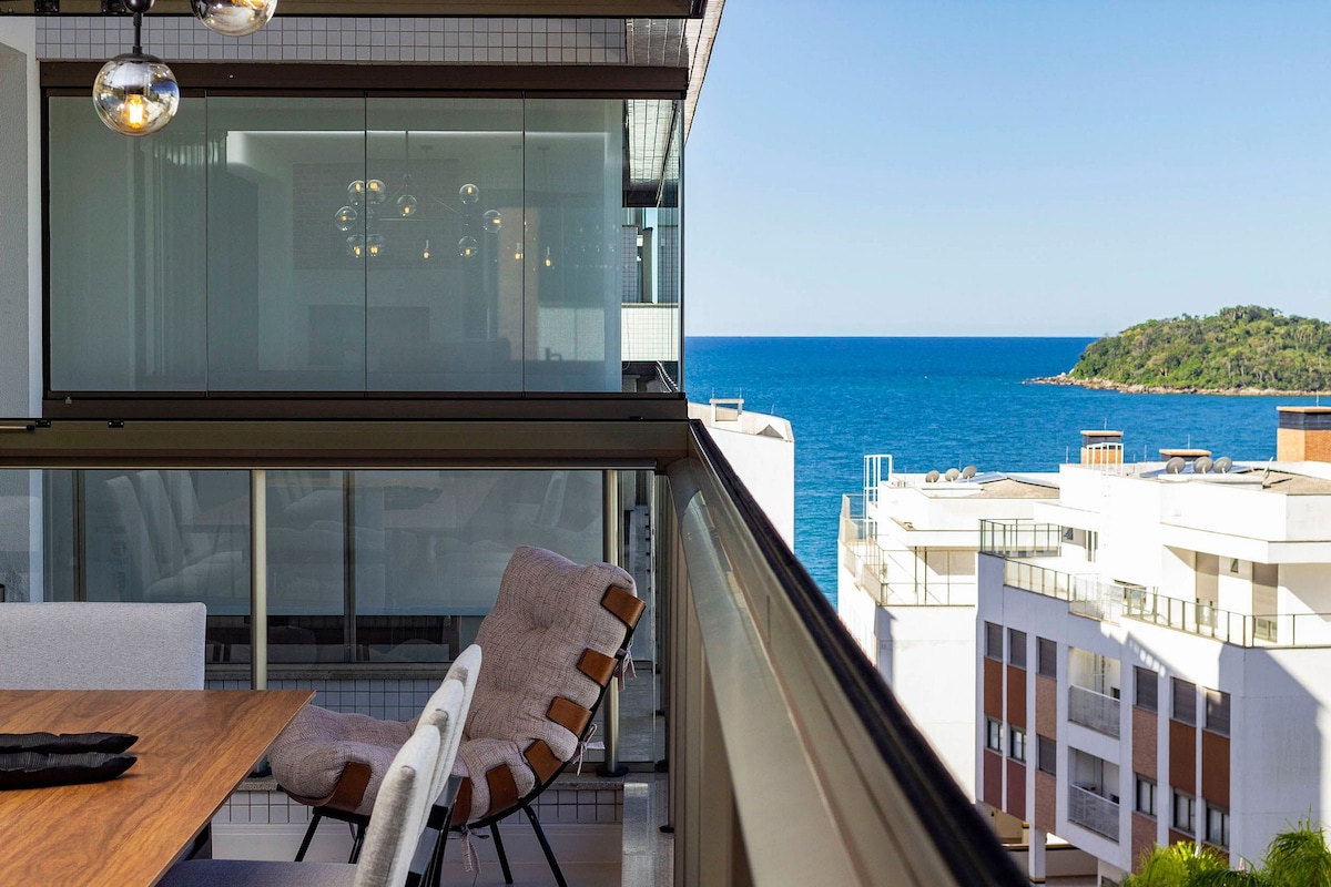 A glass-enclosed balcony offers a scenic view of the ocean and surrounding shoreline. Comfortable seating is provided alongside a wooden dining table, enhancing the space for relaxation and meals. The clean lines of the architecture contribute to a modern aesthetic.