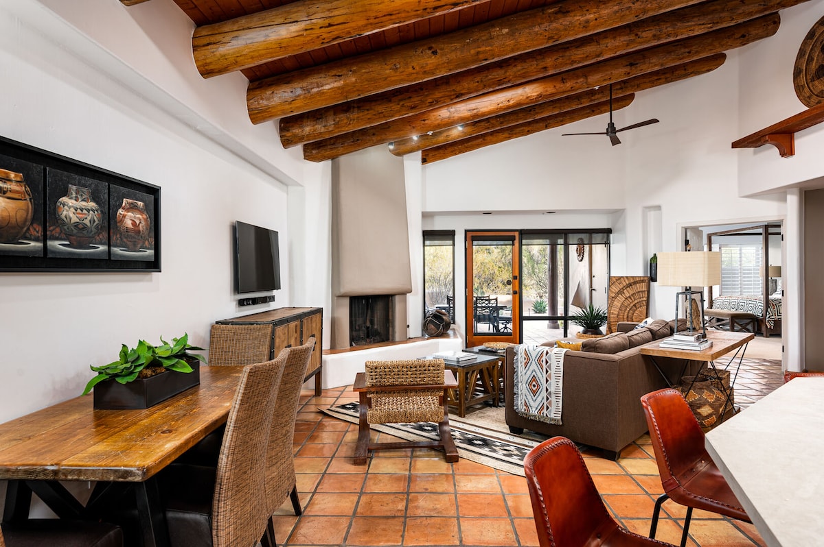 A spacious living area is revealed, featuring natural wood beams on the ceiling and terracotta tile flooring. A comfortable seating arrangement with a couch and chairs surrounds a coffee table, while a fireplace is located in the corner. A dining table with chairs is positioned nearby.