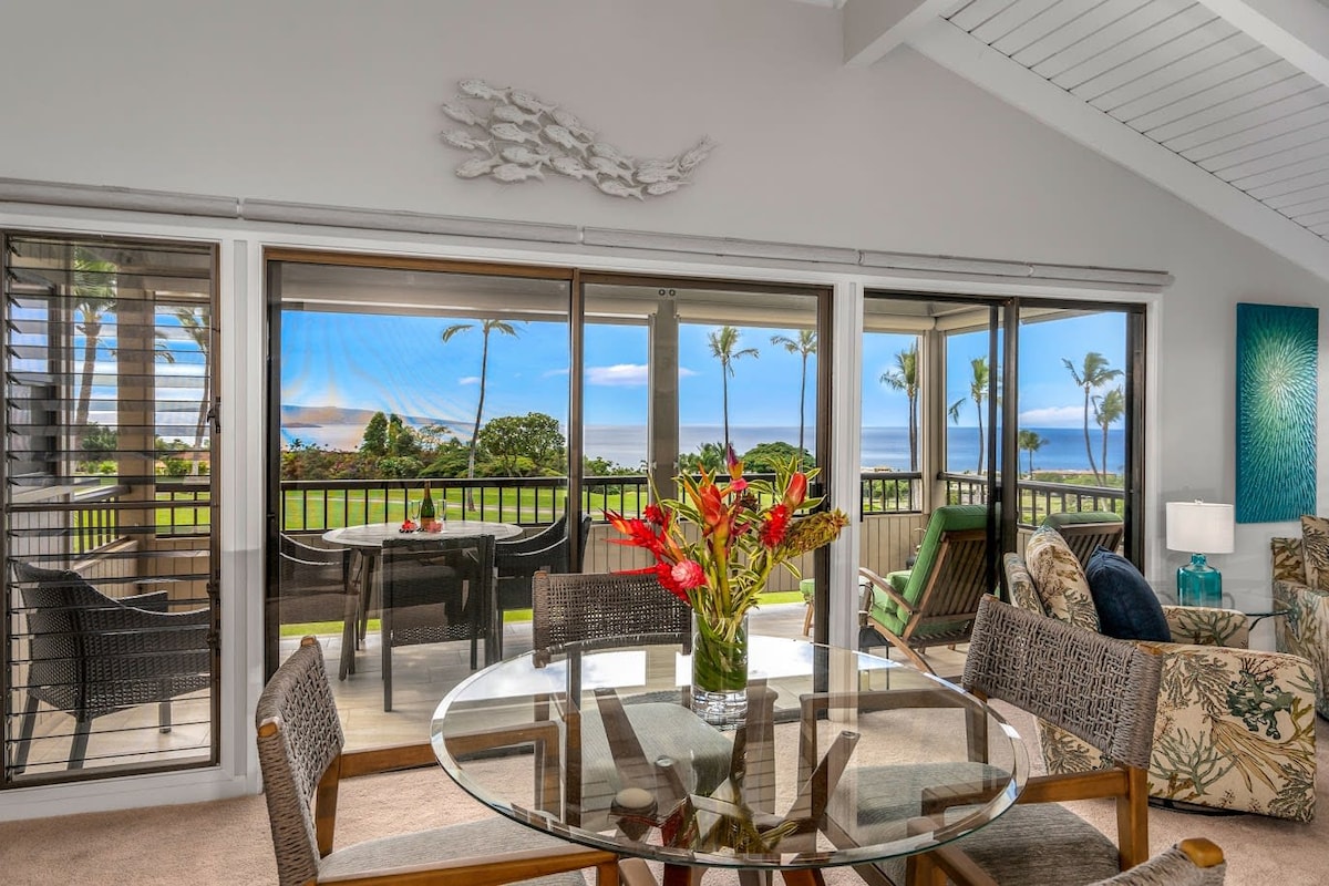 A modern dining area is visible, featuring a glass table surrounded by woven chairs. Large sliding glass doors open to a scenic lanai, showcasing ocean views and swaying palm trees. Fresh flowers add a vibrant touch to the table, enhancing the inviting atmosphere.