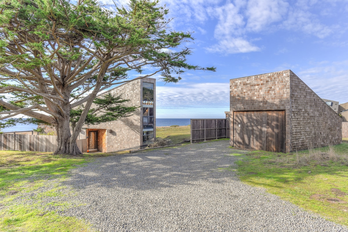 A gravel driveway leads to the modern vacation home, framed by a large tree on one side. The two structures feature a mix of wood and stone exteriors, with expansive windows reflecting the coastal landscape. The ocean is visible in the distance under a bright blue sky.