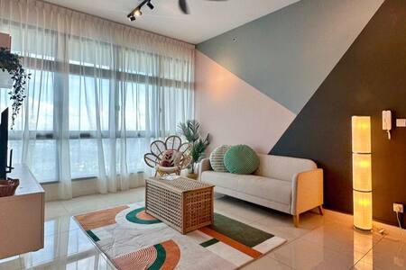 A cozy living area features a light-colored sofa and a rattan chair, positioned on a patterned rug. Large windows dressed in sheer curtains allow natural light to fill the space. A decorative floor lamp and plants add warmth to the environment.