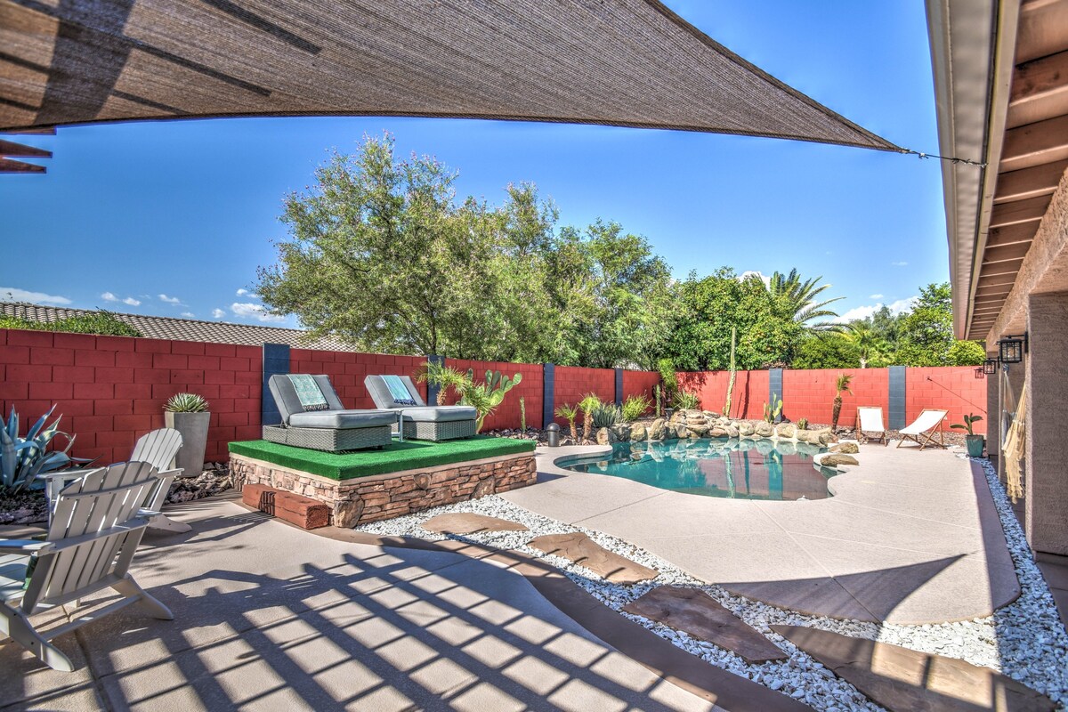 An outdoor space features a refreshing pool surrounded by desert landscaping. Two lounge chairs are positioned beside the pool, and a shaded area is provided by a large fabric canopy. The walls are adorned with vibrant red and green accents, enhancing the comfortable ambiance.