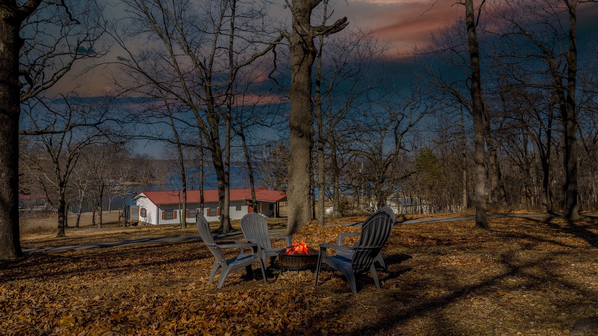 A serene outdoor space is highlighted by a fire pit surrounded by four cozy chairs. The scene is framed by tall trees and fallen leaves, with distant views of a lake under a dramatic sky.