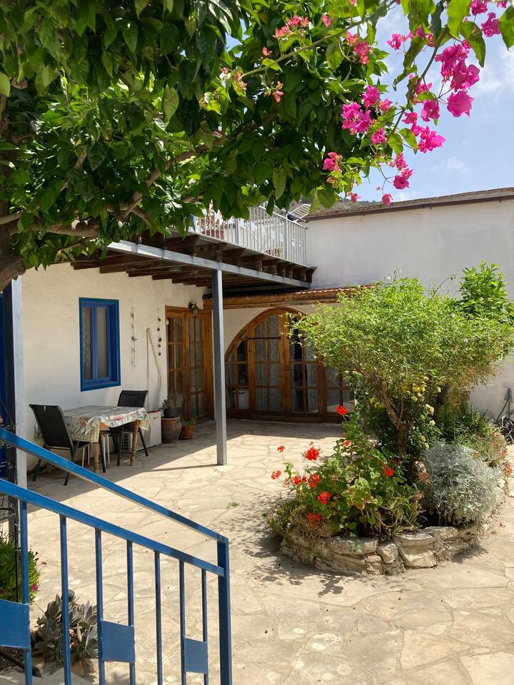 A charming courtyard is surrounded by vibrant greenery and blooming flowers. A traditional stone path leads to an entry with wooden arched doors. A dining table and chairs are placed under a shaded area, inviting outdoor meals in a tranquil setting.
