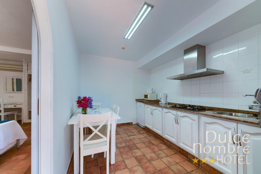 A clean, well-equipped kitchen area is seen, featuring white cabinetry and a neutral tile floor. A small dining table with two chairs is placed near a window. A gas stove and microwave are included, alongside essential kitchen appliances arranged neatly.