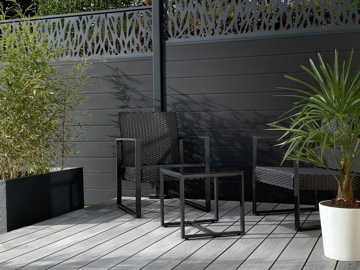 An outdoor terrace features two black wicker chairs and a small table, positioned on a wooden deck. A potted plant adds a touch of greenery, while a textured black fence provides privacy and elegant detailing.