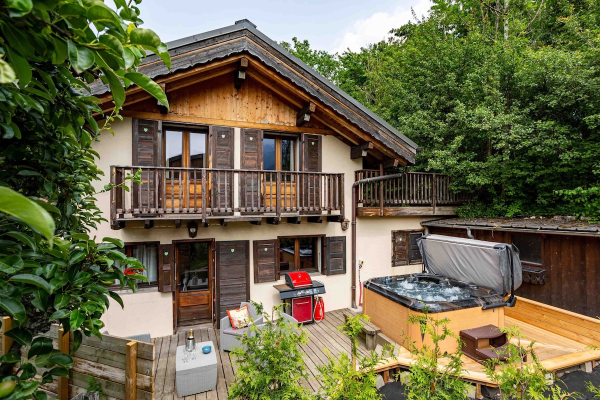 The exterior of the chalet is framed by lush greenery, featuring wooden balconies accessible from the upper level. A hot tub is positioned on the terrace, alongside a barbecue grill. Warm wood accents enhance the inviting appearance of the traditional architecture.