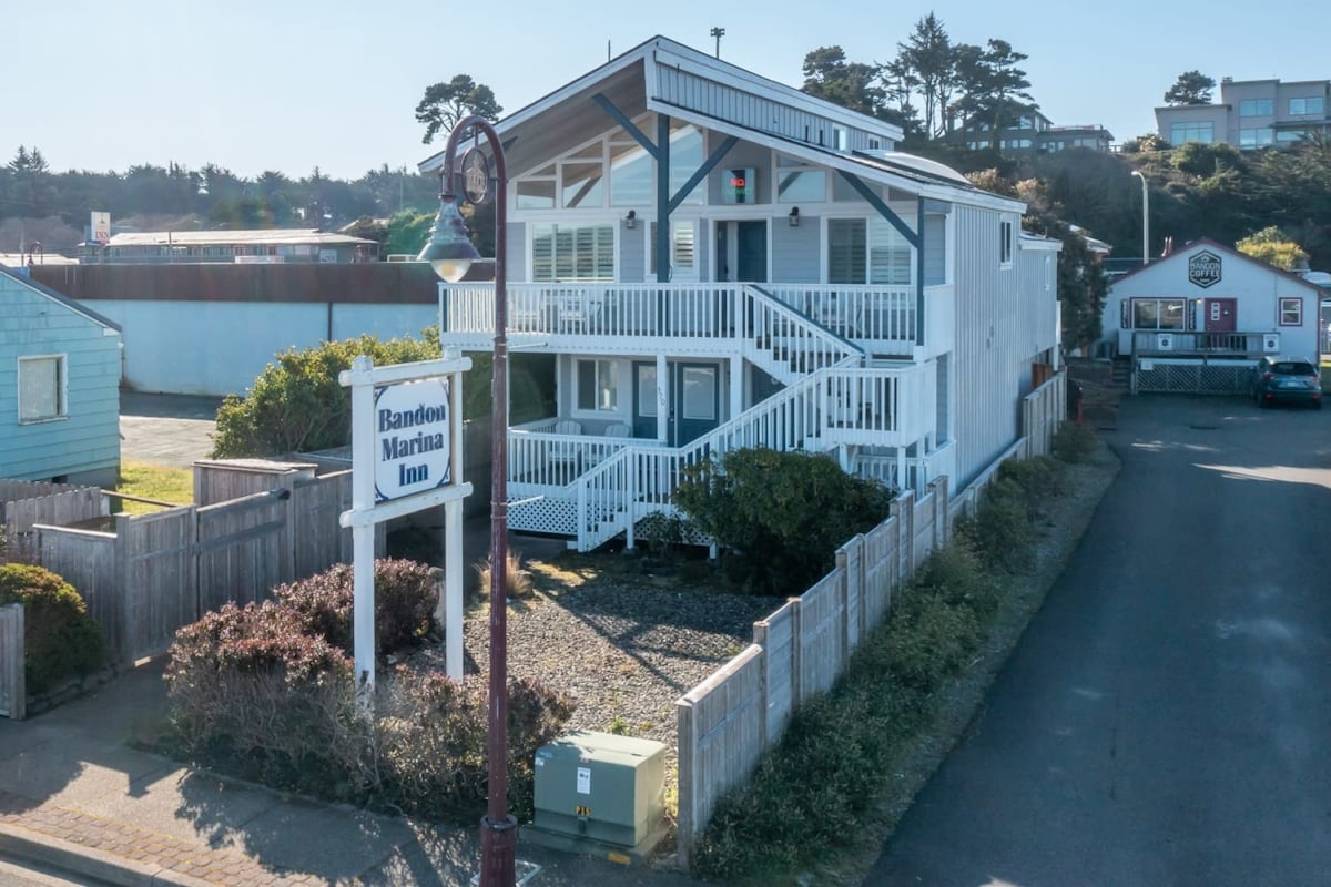 The exterior view of Bandon Marina Inn is presented, showcasing a two-story building with a white and blue façade. A sign identifying the inn is visible in the foreground, surrounded by low shrubbery and a gravel landscape. Nearby structures and coastal scenery can be seen in the background.