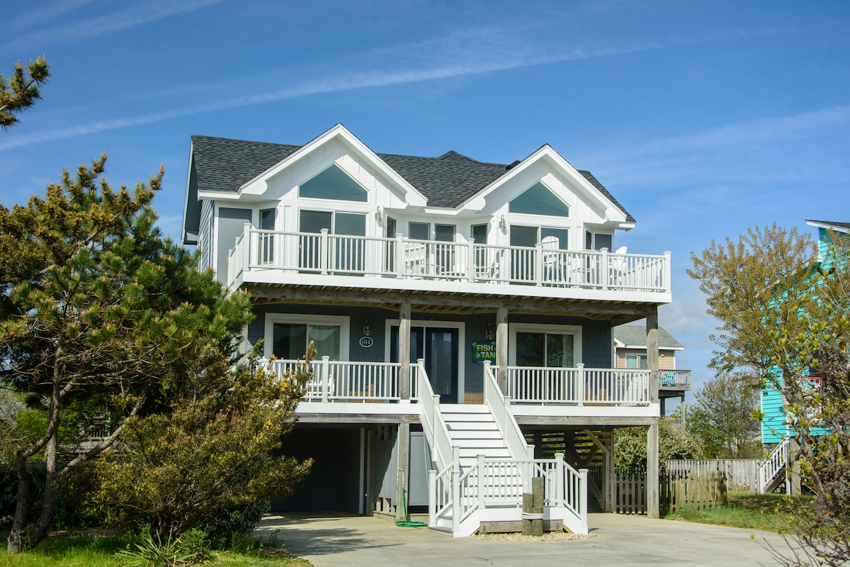 The two-story beach house features a light exterior with multiple balconies, offering ample outdoor space. Stairs ascend from the driveway to the main entrance, while the surrounding landscape includes manicured shrubs and trees, presenting a welcoming entry to the retreat.