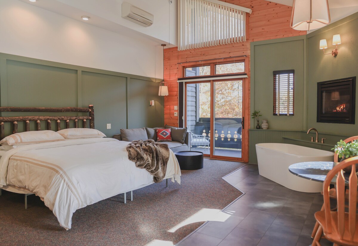 A spacious bedroom features a large king-sized bed, complemented by cozy bedding and a sitting area with a couch and a round ottoman. A freestanding soaking tub is positioned beside a gas fireplace, while large windows offer natural light and views of the outdoor deck.