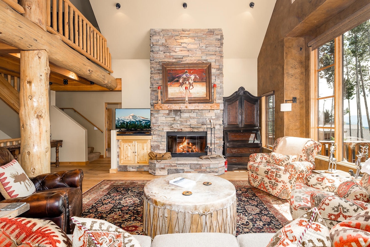 A warm living area features a stone fireplace with a television mounted above it. Colorful patterned chairs and a round coffee table surround the space, while large windows invite natural light and offer views of the surrounding trees. A wooden staircase is visible in the background.