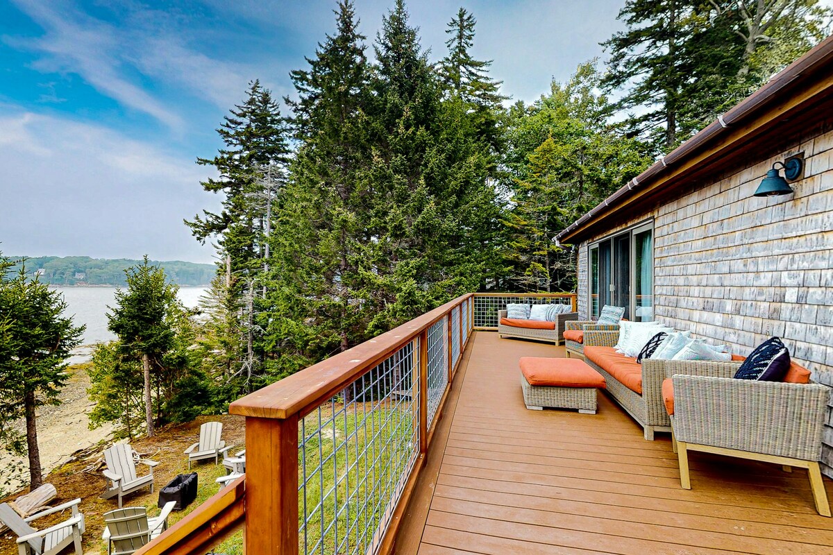 A spacious outdoor deck features comfortable seating arrangements with orange cushions, surrounded by coastal pines. The view extends toward an ocean inlet, while several Adirondack chairs are positioned along the edge, inviting relaxation in a serene natural setting.