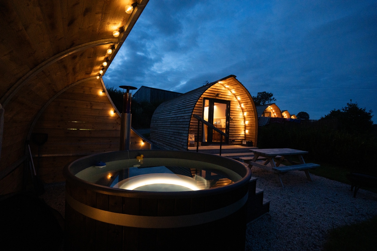 A wooden hot tub is illuminated with soft lighting, situated on a pebbled outdoor area. Cozy cabins are visible in the background, featuring wooden facades and warm lights. An outdoor picnic table is positioned nearby, enhancing the inviting atmosphere of the space.