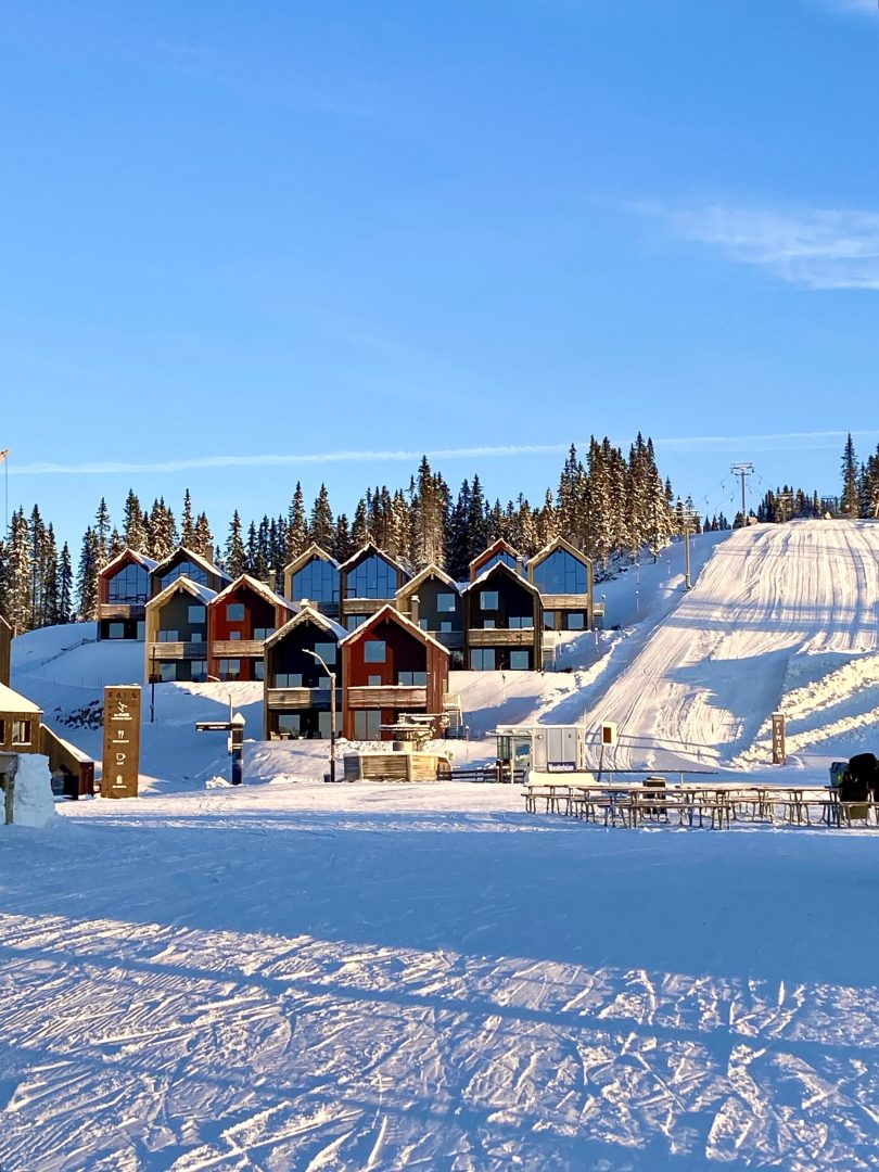 A cluster of modern multi-level buildings is set against a backdrop of snow-covered slopes and tall evergreen trees. The structures display a mix of warm colors, contrasting with the bright blue sky. A ski lift can be seen operating nearby, enhancing the winter sports atmosphere.