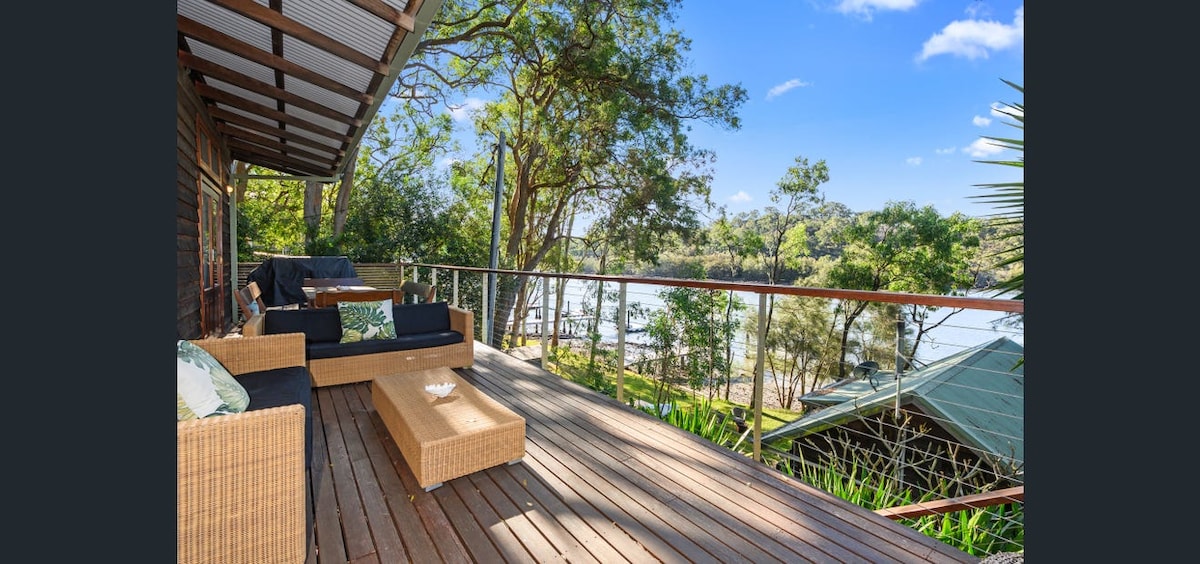 A spacious outdoor deck is seen, featuring a relaxed seating arrangement with comfortable furnishings. The deck offers expansive views of the lush greenery and the tranquil Hawkesbury River, enhancing the natural beauty of the setting.