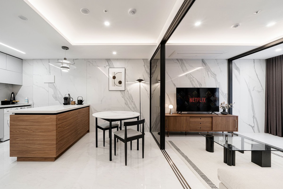 A modern open-concept living area features a sleek kitchen with minimalist cabinetry and a dining table surrounded by two black chairs. A large smart TV displays the Netflix logo, while a low coffee table rests on the polished floor, reflecting the surrounding marble walls.