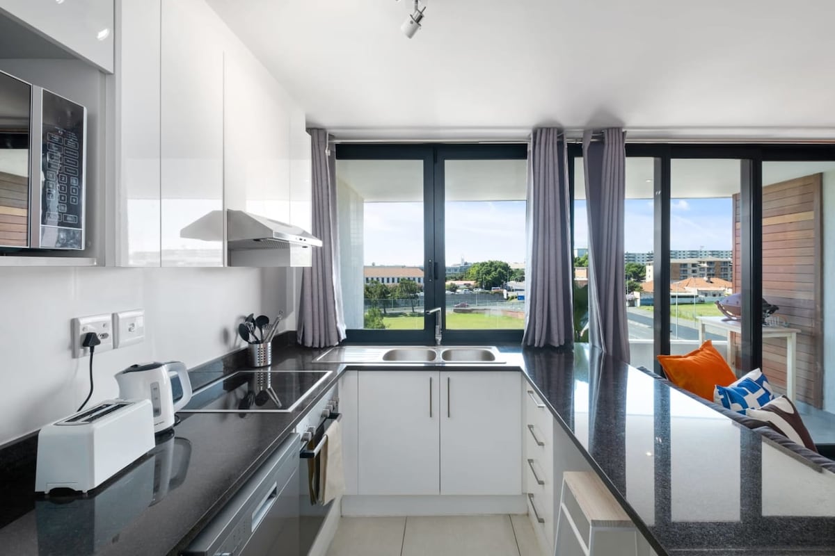 A modern kitchen features sleek white cabinetry and black countertops, equipped with essential appliances including a kettle and toaster. Large glass windows allow natural light to flood the space, with views of the outside landscape. The area is complemented by grey curtains and stylish decor.