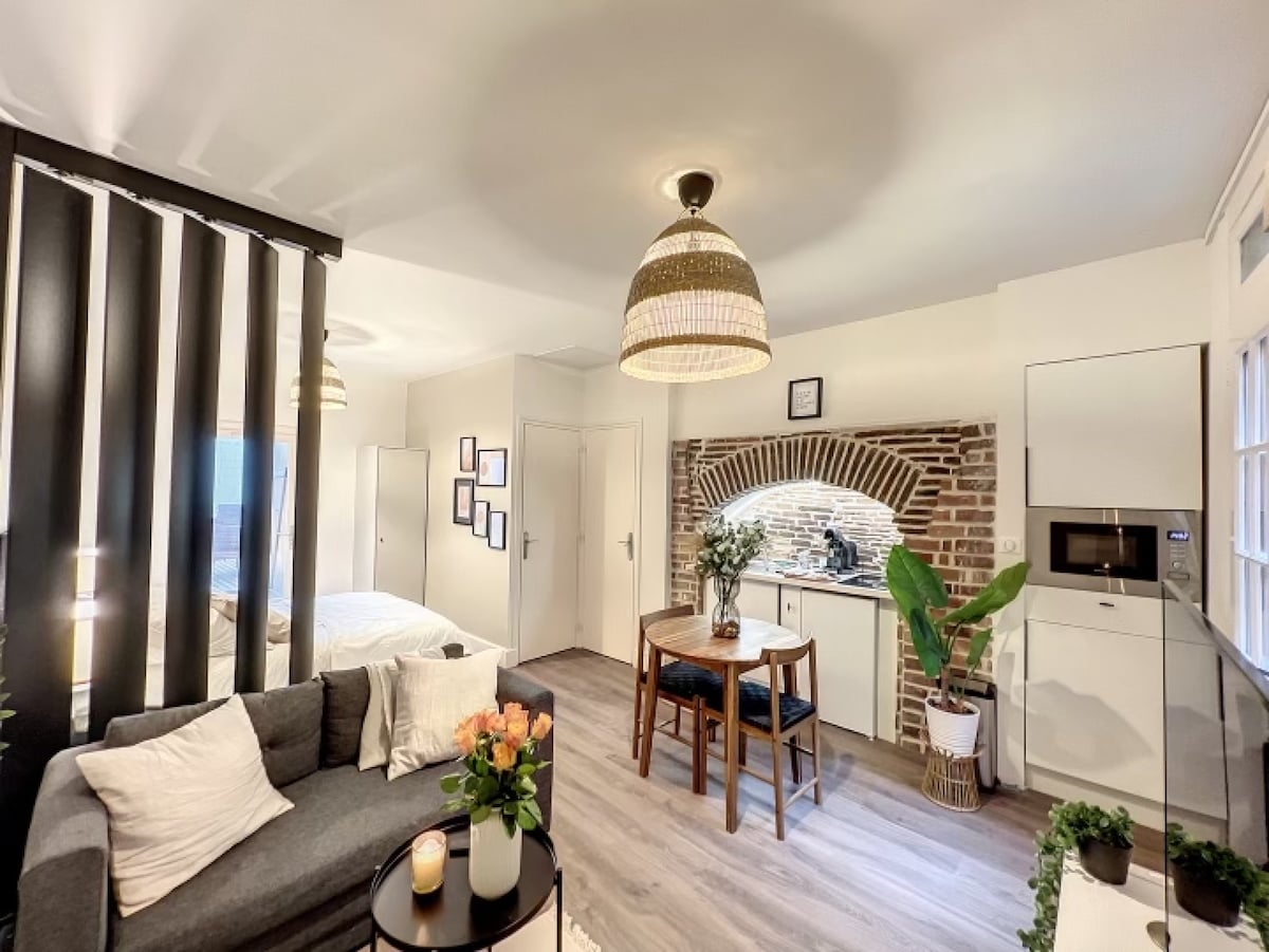 A cozy living area is highlighted by a comfortable sofa adorned with cushions. A round dining table is positioned near a rustic brick archway that leads to an equipped kitchen. Soft lighting from a pendant lamp enhances the space, complemented by greenery from potted plants.