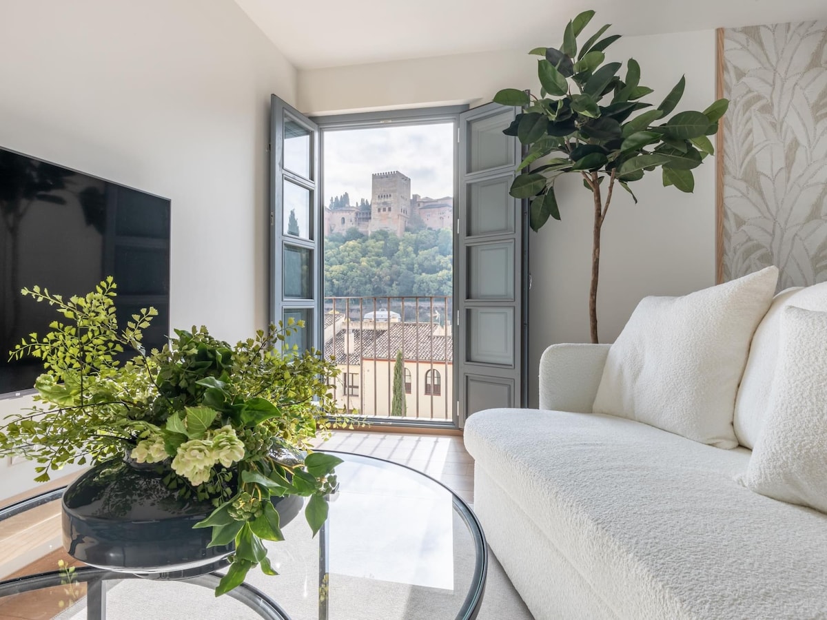 A light-filled living area is furnished with a cozy sofa and a glass coffee table accented by fresh greenery. Open doors lead to a balcony with views of a historical fortress and lush hills, enhancing the connection to the surrounding landscape.