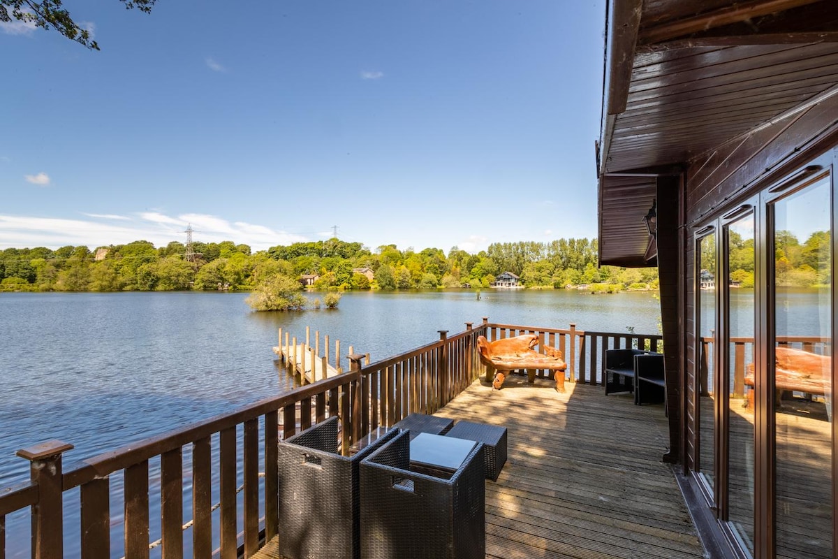 A spacious sun deck overlooks the tranquil waters of a private lake, surrounded by lush greenery. Comfortable seating is arranged around a glass table, inviting relaxation. A wooden jetty extends into the lake, providing access to the serene environment and picturesque views.