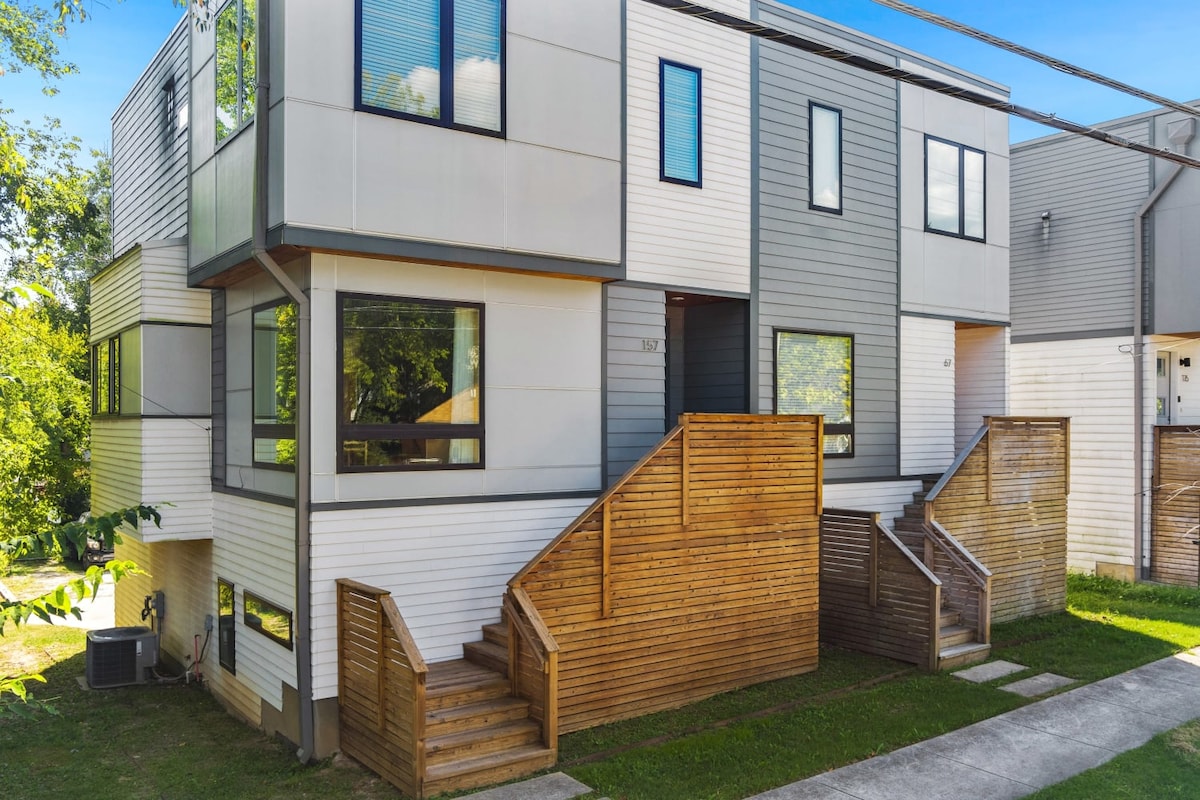 The modern townhouse features a combination of light and dark siding, with large windows allowing natural light to enter. Wooden stairs lead up to the entrance, framed by a small landscaped area. A clear pathway runs alongside the building, providing access.