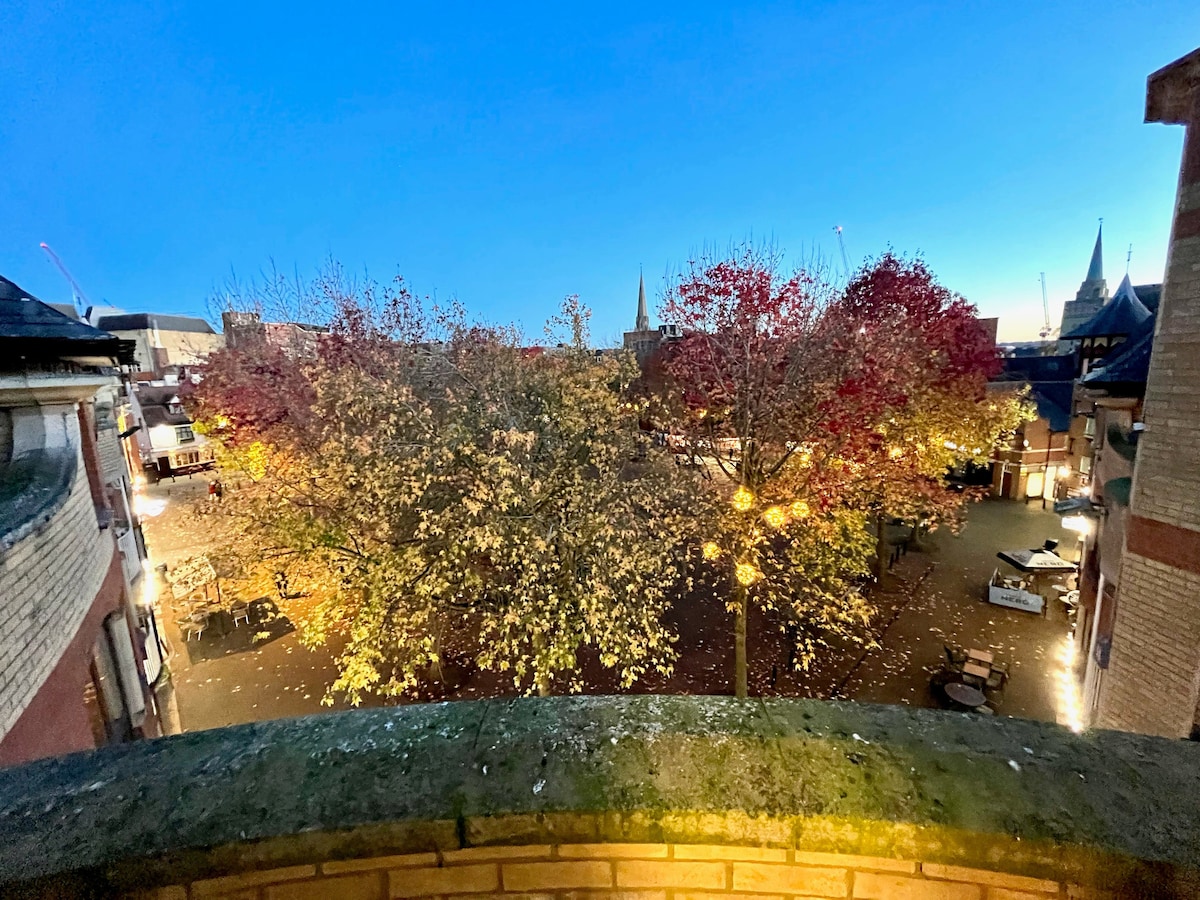 A view from the balcony captures trees displaying vibrant autumn foliage. The scene is illuminated by soft lighting, showcasing a charming square filled with fallen leaves, and historic buildings are visible in the background against the evening sky.