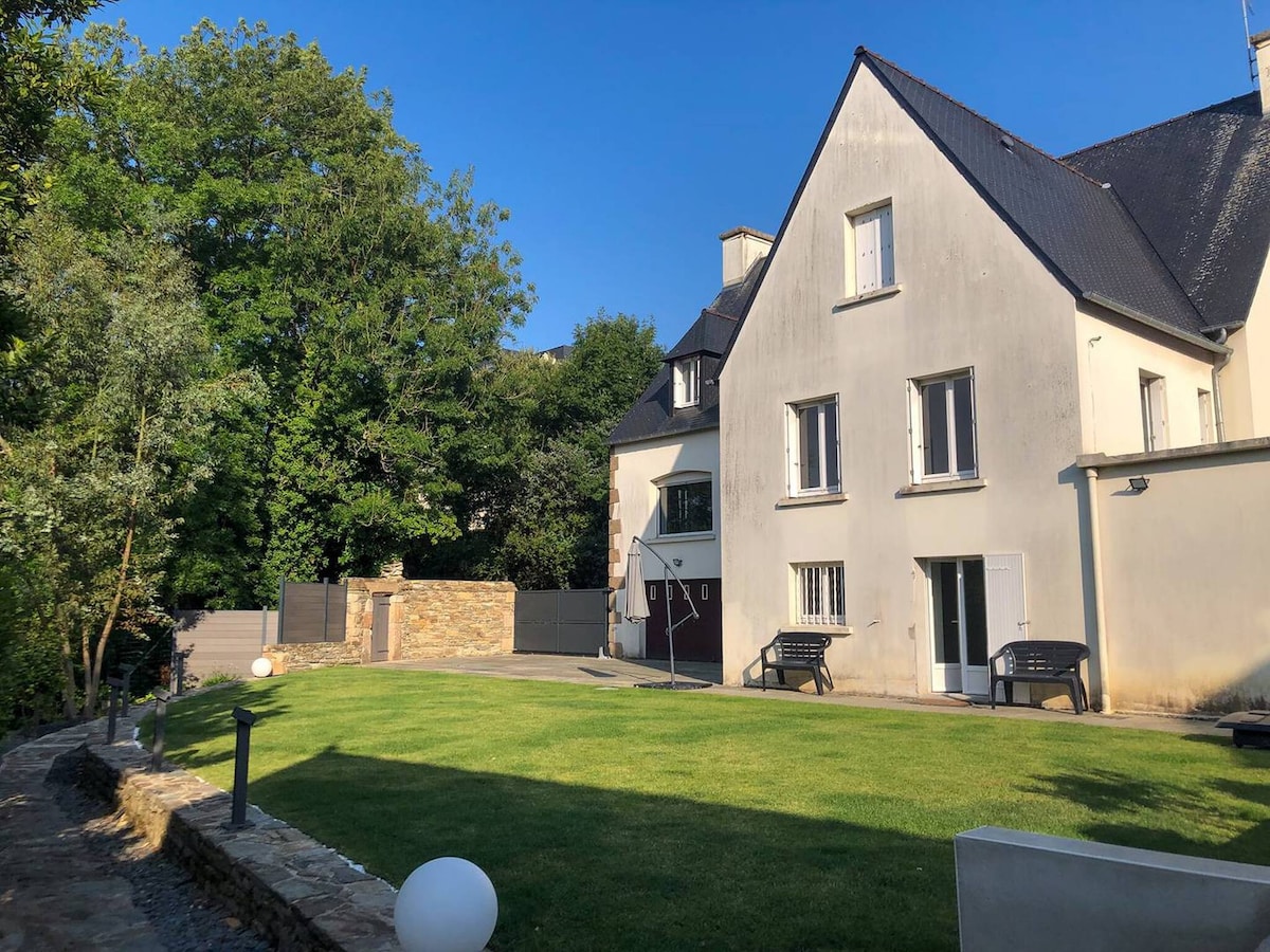 The exterior view of the house showcases a well-maintained garden with a lush green lawn and neatly trimmed hedges. The house features a traditional Breton architecture with multiple windows. Seating areas are available on the side, providing a space for outdoor relaxation.