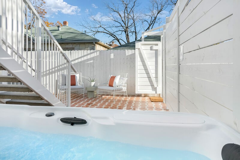 A private outdoor area is shown, featuring a hot tub filled with water, surrounded by a white wooden fence. Two lounge chairs are positioned nearby on a tiled patio, with a small table placed between them. Bright blue skies and a few trees are visible in the background.