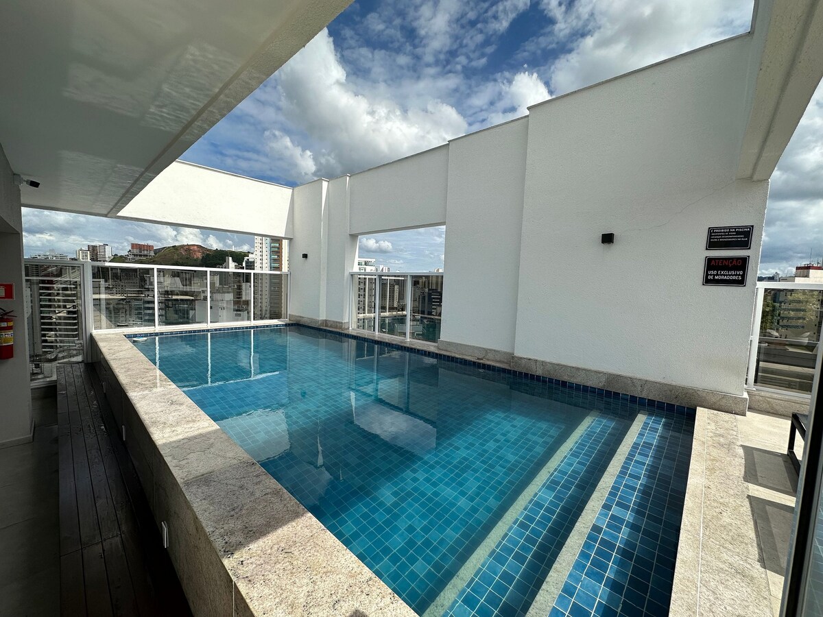 An outdoor rooftop pool features a spacious area surrounded by a modern building structure. The pool is filled with clear blue water and has light-colored stone edging. Natural light illuminates the space, and a city skyline is visible in the background.