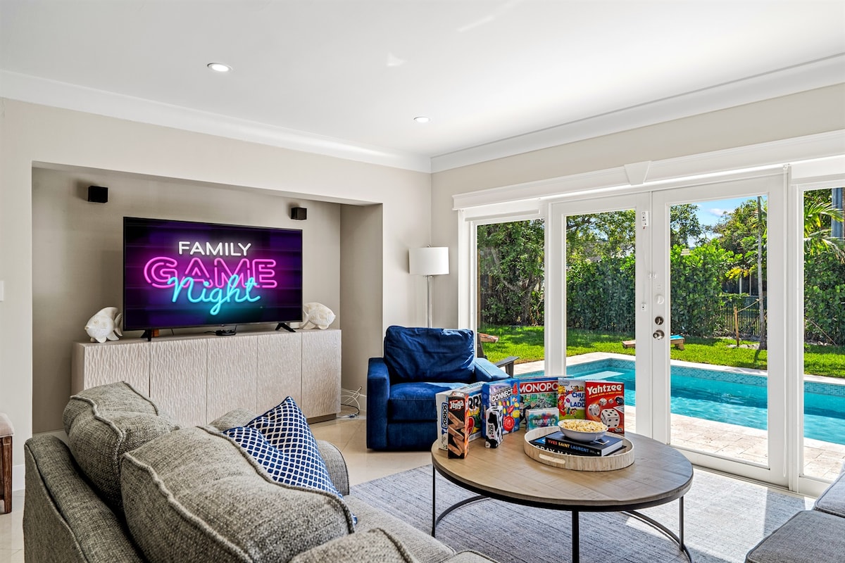 A comfortable living area features a plush gray sofa and a blue armchair, with a large flat-screen TV displaying 'Family Game Night.' A round coffee table showcases a game collection, and sliding glass doors provide views of the pool and backyard.