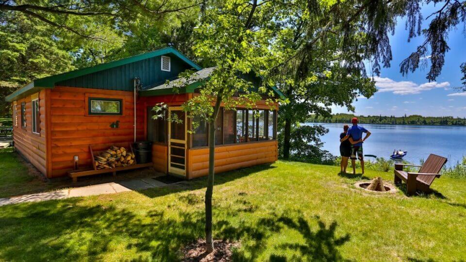 A charming cabin is framed by lush green trees, located by the serene shoreline of Big Turtle Lake. A screened porch invites relaxation, while a fire pit and seating area are positioned on the lawn, providing outdoor enjoyment with scenic lake views.