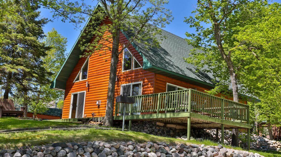 A charming lakefront cabin features a vibrant orange exterior and a green gabled roof. The structure is elevated on a grassy area, with a spacious outdoor deck overlooking stone landscaping. Large windows allow natural light to highlight the inviting facade.
