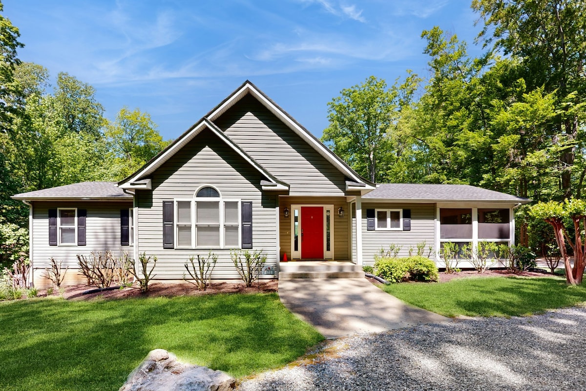 A welcoming exterior of the home features a well-maintained lawn and a gravel driveway leading to an inviting entrance. Large windows allow natural light to fill the interior, and the doorway is framed by vibrant landscaping, creating a pleasant first impression.
