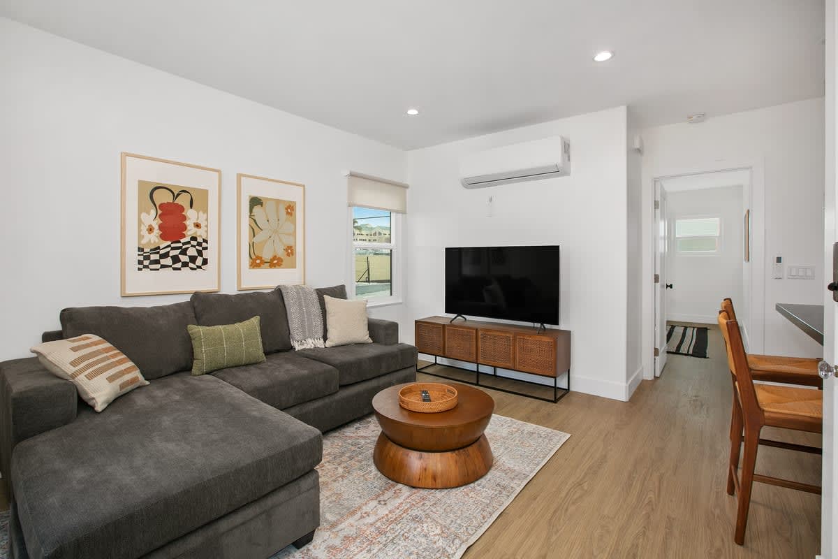 A cozy living area features a large sectional sofa adorned with decorative pillows, complemented by a wooden coffee table. Artwork in elegant frames decorates the walls. A flat-screen TV is mounted on a modern TV stand, and natural light is provided by nearby windows covered with light fabric.