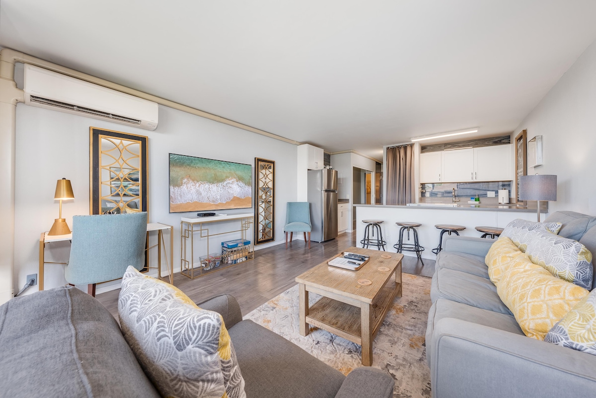 A modern living space features a comfortable sofa set with decorative cushions and a coffee table. A wall-mounted smart TV is positioned across from the seating area. The kitchen is visible in the background, showcasing sleek cabinets and stainless-steel appliances, enhancing the open-concept design.