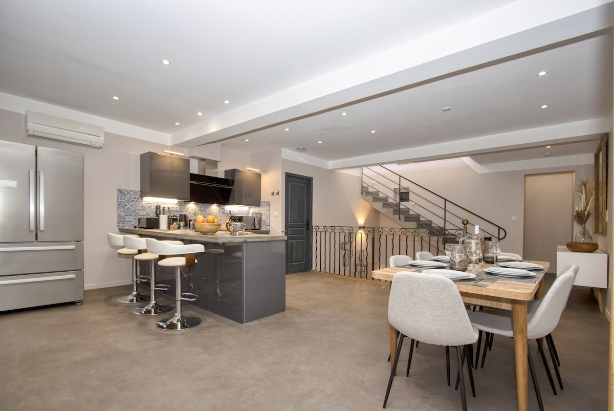 An open-plan living area is showcased, featuring modern grey cabinetry and a central kitchen island with bar seating. A cozy dining table is set for meals, surrounded by soft-toned chairs. The space is illuminated by recessed lighting and natural light from windows, enhancing its airy feel.