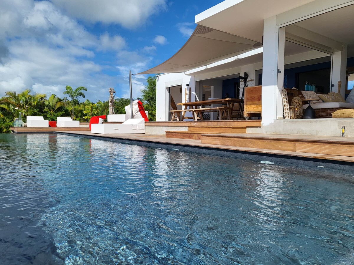 A spacious outdoor area features a sleek swimming pool with crystal-clear waters, surrounded by well-maintained landscaping. The elegant terrace includes a dining table and comfortable seating, with sun loungers positioned nearby under a large awning, providing shade and relaxation options.