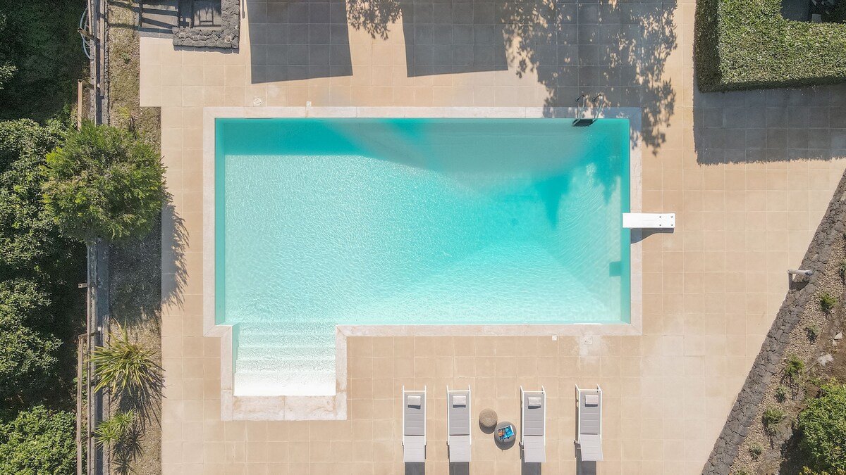 An aerial view of a rectangular swimming pool is shown, featuring clear, blue water surrounded by beige tiled flooring. The pool is accented by a diving board at the deep end and equipped with several sunbeds arranged neatly along one side.