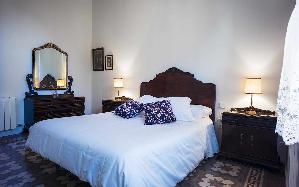 A spacious bedroom features a large bed with white bedding and decorative pillows. Antique wooden furniture is present, including a mirror and nightstands with soft lighting from lamps. The patterned tile floor adds character, creating a calming environment.