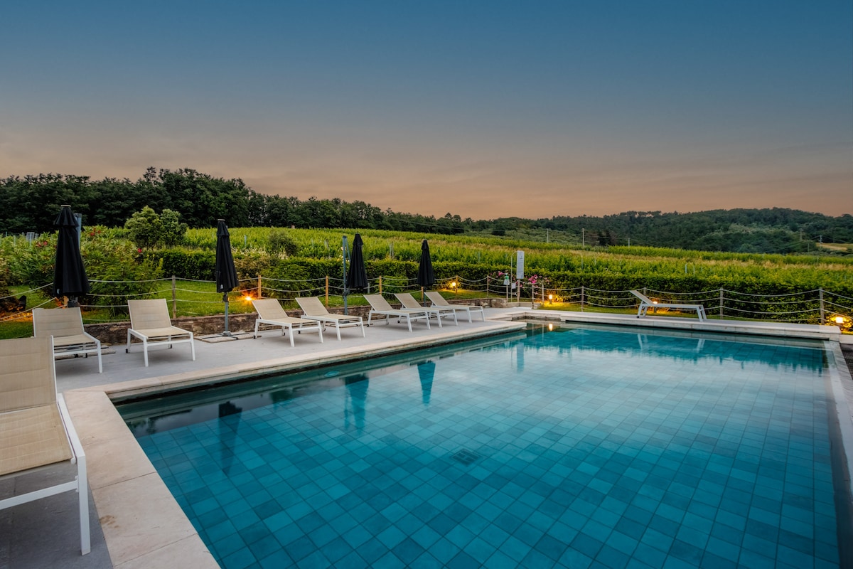 A well-maintained swimming pool is surrounded by several loungers and umbrellas. The tranquil water reflects the evening sky, while lush vineyards stretch across the background, creating a serene atmosphere complemented by soft lighting along the poolside.