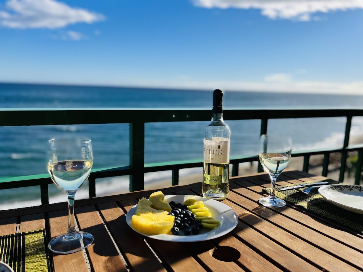 A wooden dining table on a terrace overlooks the sea, showcasing a plate of fresh fruits, including pineapple and blueberries. Two glasses of white wine and a bottle are positioned on the table, with clear blue skies and ocean waves visible in the background.