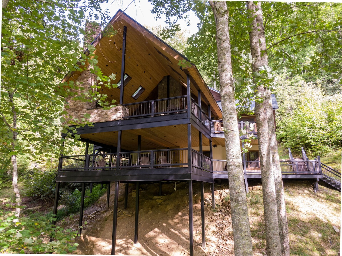 The mountain home is set among vibrant trees, featuring a multi-level structure with expansive decks. Dark wooden railings and a sloped roof enhance its modern aesthetic. A staircase leads up to the entrance, surrounded by the natural landscape.