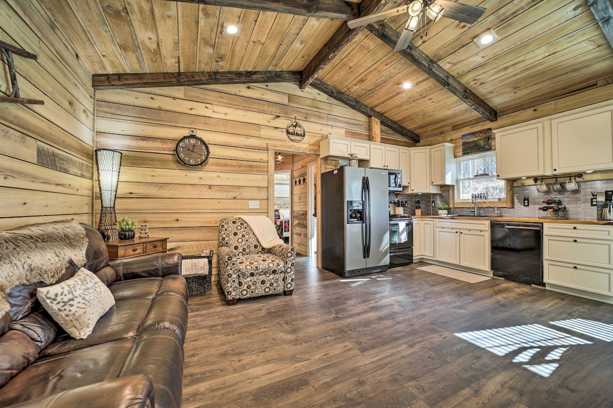 The open living space features wooden walls and ceiling beams, creating a rustic ambiance. A leather sofa and an accent chair provide comfortable seating. The kitchen is equipped with modern appliances, including a refrigerator and oven, situated against a backdrop of light cabinetry.