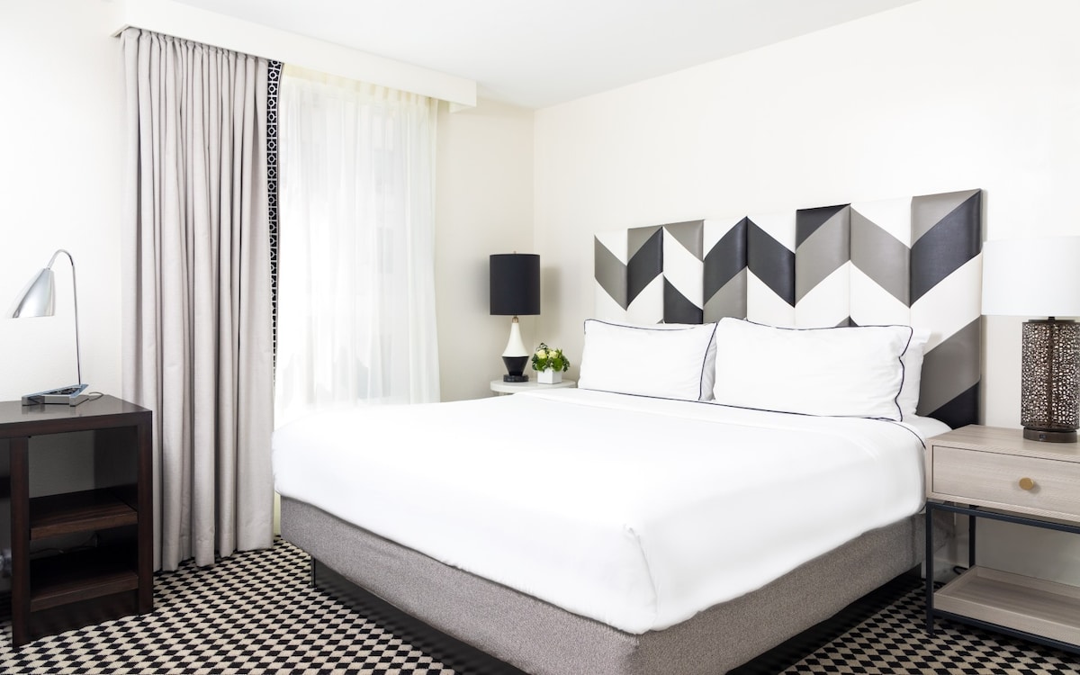 A modern bedroom features a spacious bed with a geometric black and white headboard. Light enters through a large window covered by sheer curtains. Two bedside tables hold decorative lamps, complementing the overall neutral palette of the room.