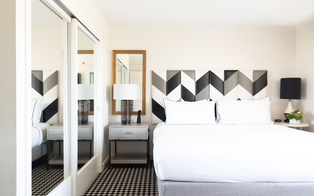 A spacious bedroom features a king bed with crisp white linens and a geometric patterned headboard in black and white. A large mirror is placed above a modern nightstand, while a stylish lamp and a small plant add to the decor.