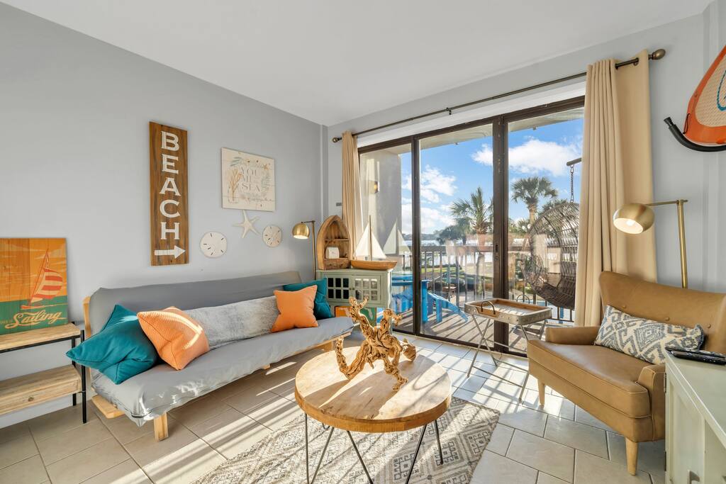 A cozy living area features a light-colored sofa with colorful cushions and a wooden coffee table. Large glass doors allow natural light to fill the space while providing views of palm trees and outdoor seating on the balcony.