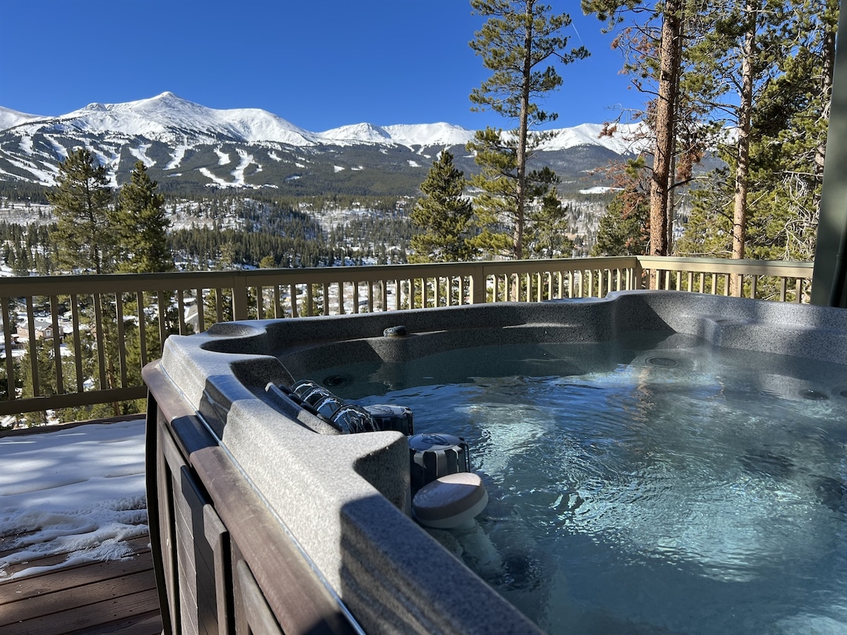 A spacious private hot tub is situated on a deck, offering stunning views of the snow-covered mountains and pine trees. The clear blue sky adds to the serene atmosphere, making it an inviting spot for relaxation.