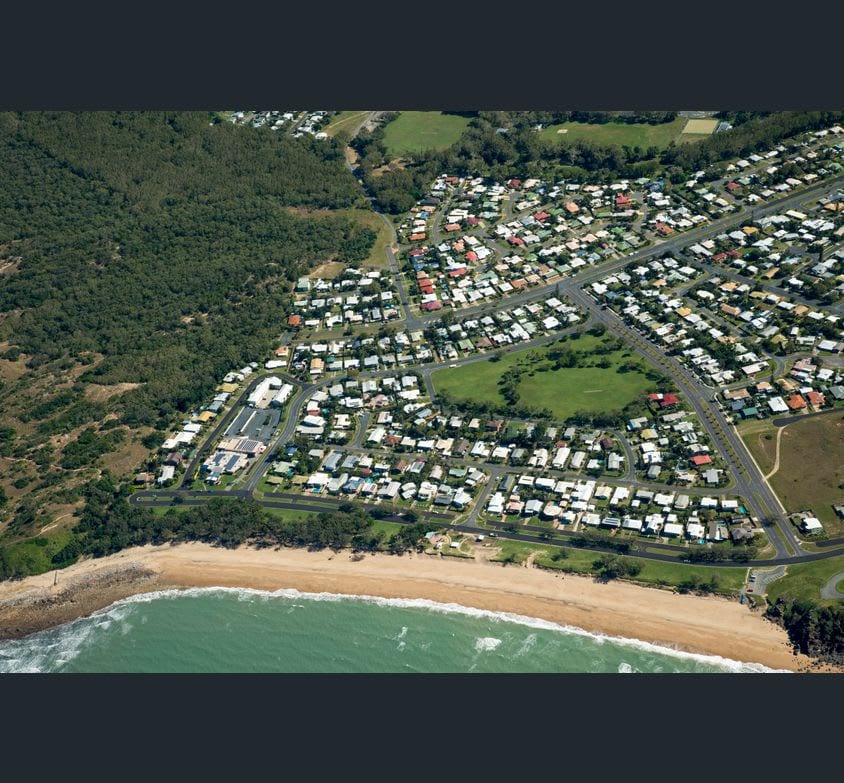 An aerial view captures a coastal community featuring a sandy beach alongside a cluster of suburban homes. Lush greenery surrounds the area, with winding roads intersecting the residential layout, providing a sense of tranquility near the shoreline.