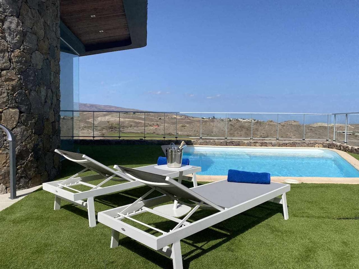 Two lounge chairs are positioned alongside a private pool, situated on a grass area. A small table rests between the chairs, holding a bucket with refreshments. The clear sky provides a backdrop to the scenic landscape visible beyond the pool.