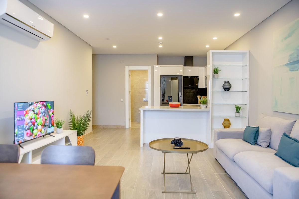 The living area features a light-colored sofa with accent pillows, a coffee table, and a Smart TV displaying colorful imagery. A dining table for four is visible to the side, while the kitchen and entry areas show modern design elements and neutral tones.